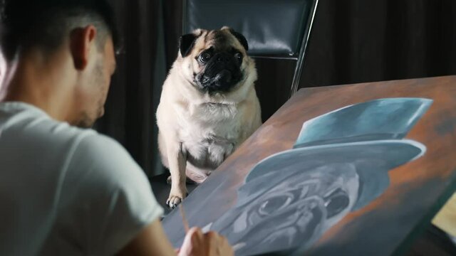 Cute pug dog posing for the artist, man painting a picture of a dog dressed in hat