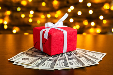 Red gift box on the USA 100 dollar bills on blurred lights background. American dollar. Christmas and New Year money present. Gift cost. Hundred. Finance and business concept. Christmas shopping