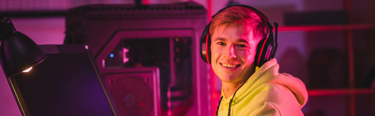 Side view of smiling player in headphones looking at camera near computer monitor and lamp, banner