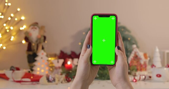 A woman using an iPhone 12 with a green screen on a Christmas background. Vertical orientation. POV. Keyed Chroma key.