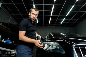 Obraz na płótnie Canvas Car detailing series, polishing concept. Professional male auto service worker, wearing black t-shirt and pants, waxing and polishing headlight of black car with polish machine, looking at camera