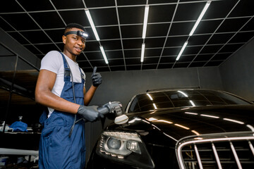 Smiling handsome young black man, professional worker of car detailing service, polishing black car with polishing machine and wax, looking at camera and showing his thumb up. Car polishing concept