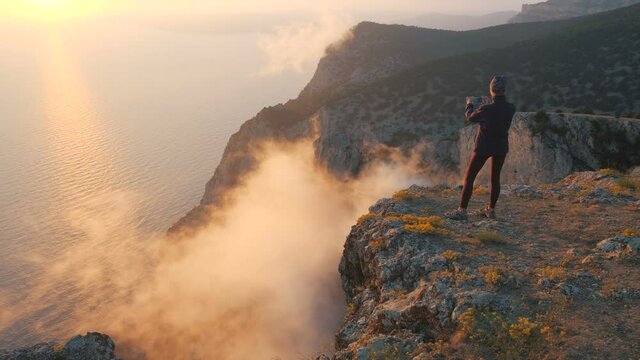 Circular shooting tourist girl standing over colorful clouds on the edge of the mountain above the sea taking pictures with smartphone. A woman at a cliff is filming beautiful sunset and Evaporation