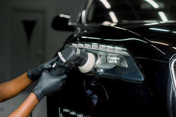Car wax polishing process. Cropped close up image of hands of male professional African worker in protective gloves, with orbital polisher in auto repair shop, polishing car headlight