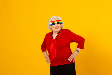 Studio portrait of a senior woman wearing a red shirt and rainbow sunglasses against a yellow background