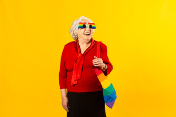 Studio portrait of a senior woman wearing a red shirt, rainbow sunglasses and a bag, against a yellow background