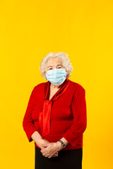 Studio portrait of a senior woman wearing a red shirt and a surgical facial mask, against a yellow background