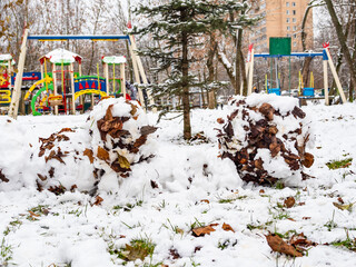 broken wall of snow fortress shaped from snowballs made from the first snow with fallen leaves on lawn of children playground in city on cold autumn day