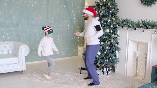 Dad dances with his little son while enjoying Christmas together in an ornate room at home with brightly lit lights and a Christmas tree. Fun activity with child, hobby, new year winter holidays celeb
