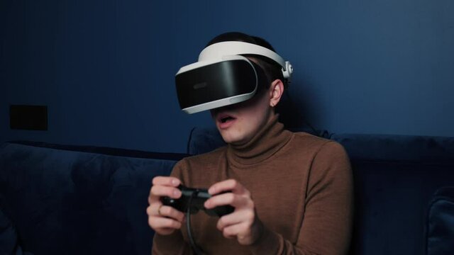 Attractive scared caucasian man hold joystick and wear virtual reality glasses headset helmet on sofa at home. Scared gamer playing game using VR glasses at home.