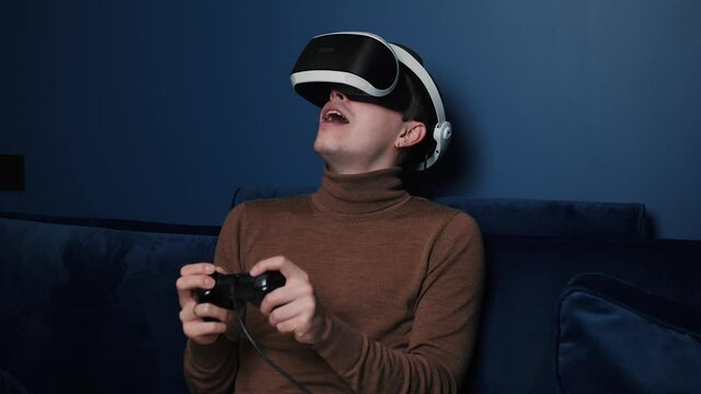 Attractive caucasian man hold joystick and wear virtual reality glasses headset helmet on sofa at home. Happiness gamer playing game using VR glasses at home.
