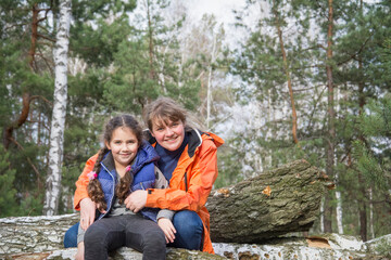On a bright spring day, mom and daughter are joyful sitting on a log in the forest.