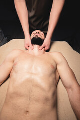 Male hands of a masseur doing neck massage to a bearded male athlete in a dark room of the massage room