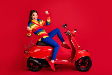 Fototapeta na wymiar Photo portrait of excited girl celebrating with raised fists leaning back sitting on scooter isolated on vivid red colored background