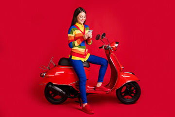 Fototapeta na wymiar Photo portrait of woman texting holding phone in two hands sitting on scooter isolated on vivid red colored background