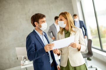 Man and woman with protective facial masks discussing with paper in hands indoors in the office with young people works behind them