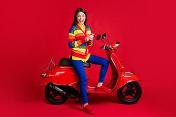 Plakat Photo portrait of excited girl holding phone in two hands sitting on scooter isolated on vivid red colored background