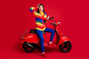 Fototapeta na wymiar Photo portrait of shocked girl touching head holding phone in one hand sitting on scooter isolated on vivid red colored background