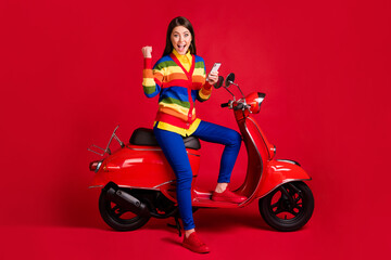 Fototapeta na wymiar Photo portrait of girl celebrating raising one fist holding phone in hand sitting on scooter isolated on vivid red colored background