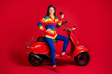 Plakat Photo portrait of confident woman holding plastic card in one hand sitting on scooter isolated on vivid red colored background