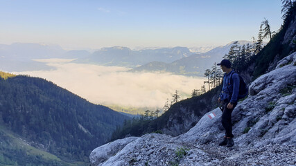 A man with a hiking backpack enjoying the view on the city below, while hiking to the top of Grimming in Austria. The valley in shrouded in fog. Early morning in the Alps. Expedition