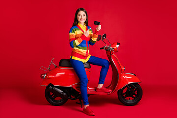 Fototapeta na wymiar Photo portrait of woman pointing finger at credit card holding in one hand sitting on scooter isolated on vivid red colored background