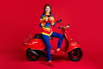 Plakat Photo portrait of girl holding credit card in two hands sitting on scooter isolated on vivid red colored background