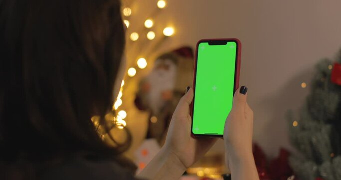 Back view of woman holding iPhone. Green screen. Woman talking, smiling, laughing in a video calling with her family during Christmas time. Christmas quarantine at home. Dolly shot