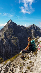 A woman sitting at the edge of a mountain with a view on a vast valley. There are sharp mountains and high peaks around. The Alpine slopes are almost barren. Lush green valley. Bright day. Freedom.