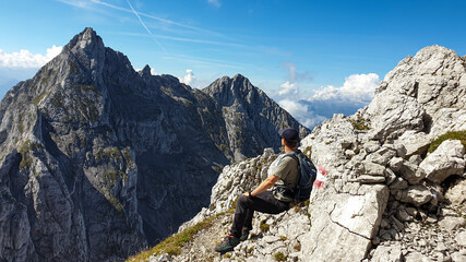 A man sitting at the edge of a mountain with a view on a vast valley. There are sharp mountains and high peaks around. The Alpine slopes are almost barren. Lush green valley. Bright day. Freedom.