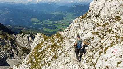 Fototapeta premium A man walking on a very narrow and steep pathway along a mountain wall in Austrian Alp, leading to the top of Grimming. There is a small city in the bottom of the valley. Dangerous trail. Adventure