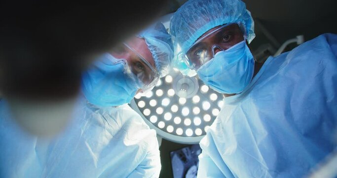 Plastic surgery. Mixed-races male specialists in medical uniform, goggles and mask on face performing operation with medical tools. Teamwork of surgeons in the operating room in hospital. Close up