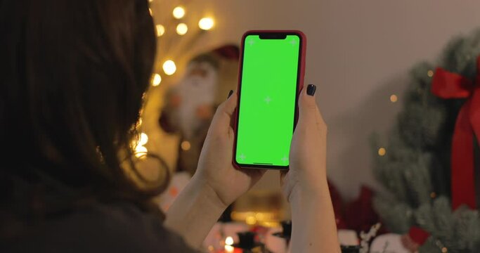 Back view of woman holding iPhone. Green screen. Woman talking, smiling, laughing in a video calling with her family during Christmas time. Christmas quarantine at home.