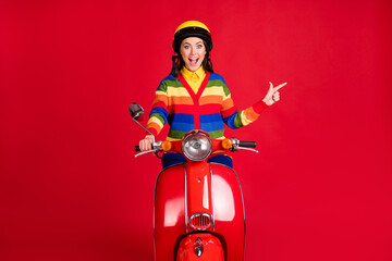 Photo portrait of screaming girl pointing finger to side driving retro scooter isolated on vivid red colored background