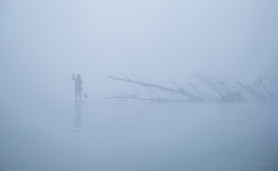 unrecognizable female with paddle on the sup board on the river surrounded of dense fog near tree