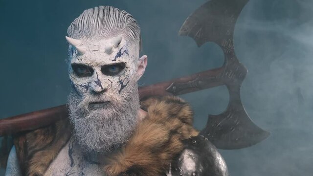 Risen undead warrior with horns and frosty skin poses in smokey atmospheric background holding huge double axe on his shoulder.