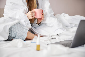 Young woman with flu drinking tea and using laptop at home