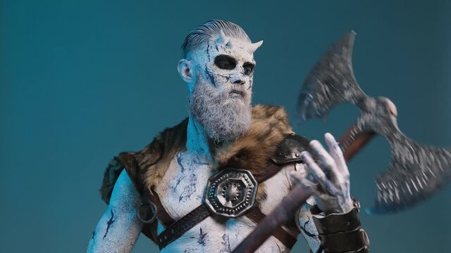 Mythical evil undead barbarian with horns and frosty skin puts his huge double axe in front of himself and staring at camera with blue eyes in dark blue background.