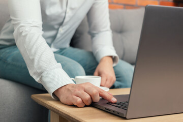 Unrecognized man, businessman or freelancer sitting on sofa and working at laptop on table with coffee cup, work from home