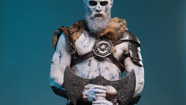 Evil and fantasy portrait of a undead warrior with pale skin and light armour posing in blue background holding his huge double axe in front of himself and looking at camera.