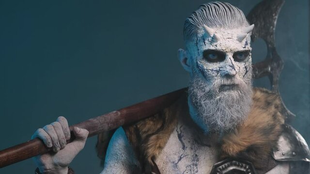 Risen undead warrior with horns and frosty skin poses in smokey atmospheric background holding huge double axe on his shoulder.
