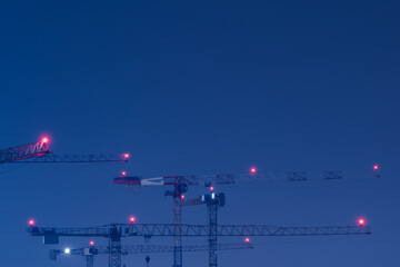 Looking up at group of tall and high tower cranes with red position lights at large construction site for housebuilding at night with blue night sky and copy space