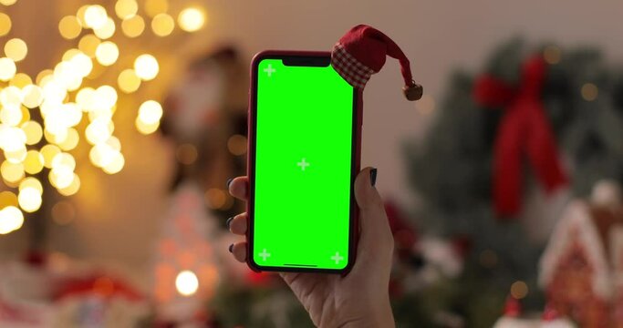 Woman sitting on chair and watching iPhone Xs during Christmas eve. Green screen display. Remote video call for Christmas. Relaxing at home during Christmas holidays.