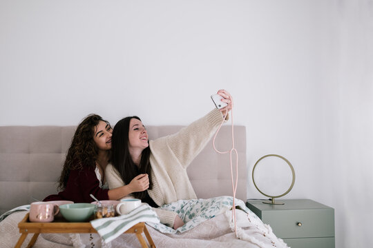Two white girls with brown hair taking a selfie in bed. They wear lingerie and a cardigan