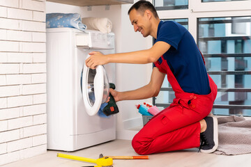 Plumber in overalls with tools is repairing a washing machine in the house