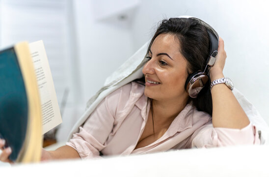 Beautiful Smiling Woman Lying On Bed With Blanket Over Head While Reading Book And Listening Music