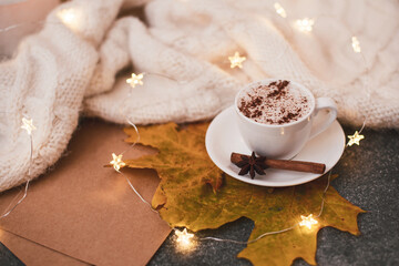 A cup of cappuccino near a warm beige blanket, dried maple leaf, a garland.
