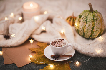 Сomposition in brown-beige colors in the hygge style. Cozy knitted blanket, candle flame, garland, small pumpkins, a cup of hot cappuccino with spices.