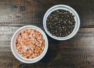 Pink salt and peppercorns in white ceramic bowls on a wood table 