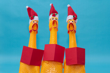 Three rubber chickens with Santa Claus hats and red books singing Christmas carols on blue...